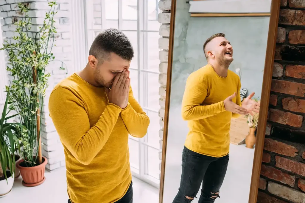 Man standing beside a mirror, appearing distressed, while his reflection looks joyful.