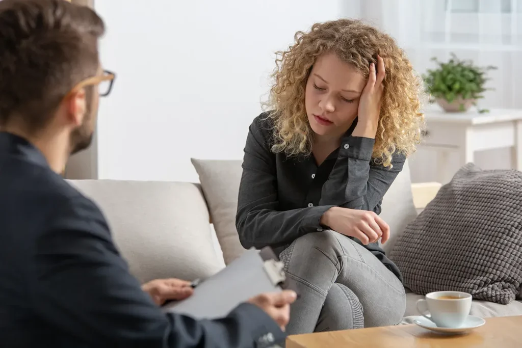 woman sitting on a couch, looking distressed, speaking with a therapist holding a clipboard.