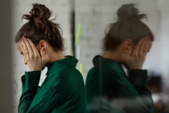 A distressed woman with her hair in a messy bun covering her face in her hands, next to a reflection of herself seen through a glass window pane.