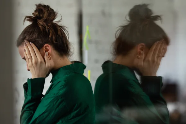 A distressed woman with her hair in a messy bun covering her face in her hands, next to a reflection of herself seen through a glass window pane.