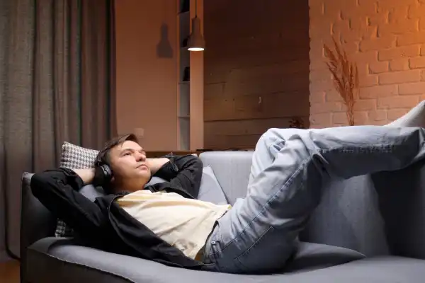 A young man reclining on a gray sofa in a warm, cozy room while listening to music with over-ear headphones.