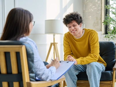 A young man in Yellow smiling while speaking with a counselor during an in-person talk therapy session.